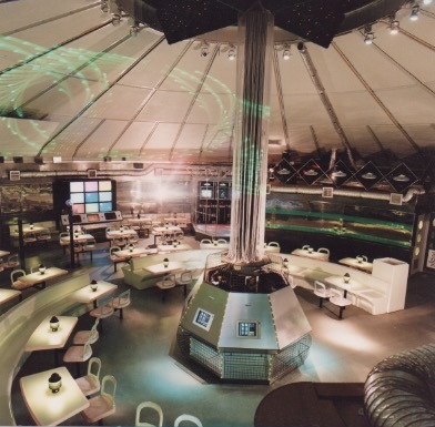 Overhead view of Megatron interior during laser show. Central computer console, bank of TV screens, and dining tables all visible.