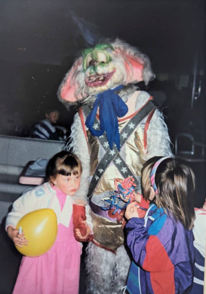 Ragged-looking Fidjet mascot stands with children inside Megatron. One girl holds his hand while holding a balloon in her other hand.